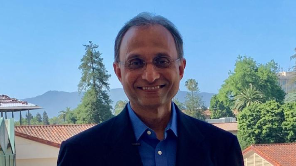 A smiling man with glasses, wearing a blue, collared shirt and a suit jacket.