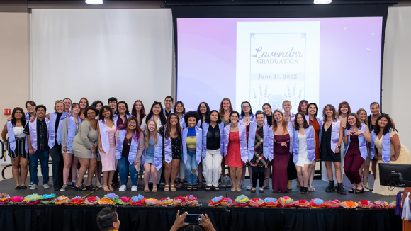 A group of students wearing light purple commencement stoles in front of a screen that says