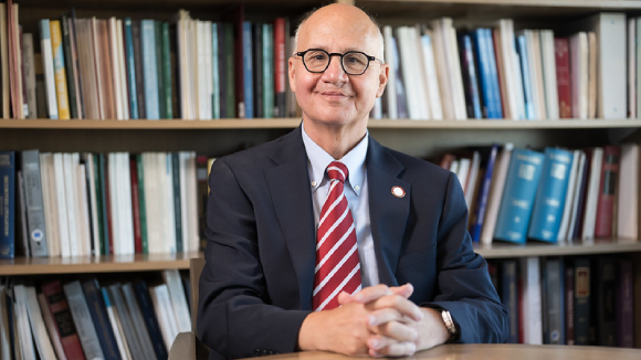 A smiling man in a suit and tie, sitting in front of a bookshelf filled with books.
