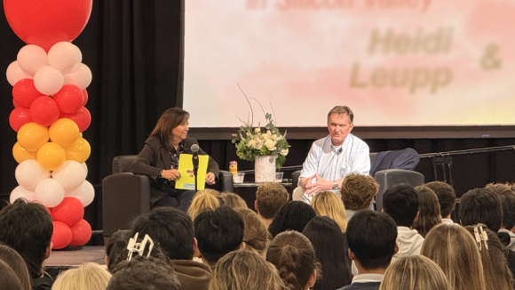 A man and woman sit in armchairs on a stage, in front of an audience, in the middle of a conversation.