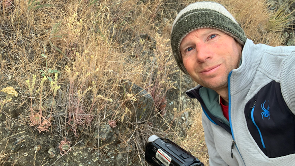 A man in a knit beanie, holding a camera and crouching in front of a wild succulent.