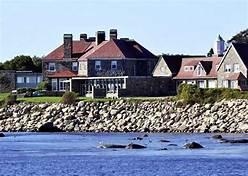 Large house with red roof near rocky shoreline and bright blue water.