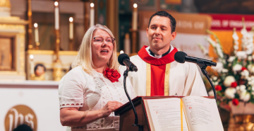 A woman in businesswear speaks at a podium in a church. Standing at her left shoulder is a man in clerical collar and robes. 