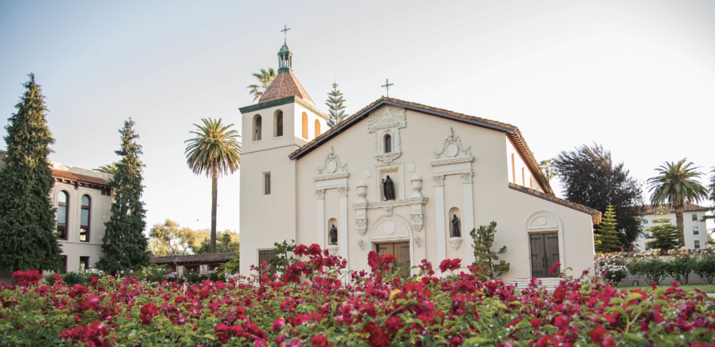 A white mission church surrounded by red flowers. 