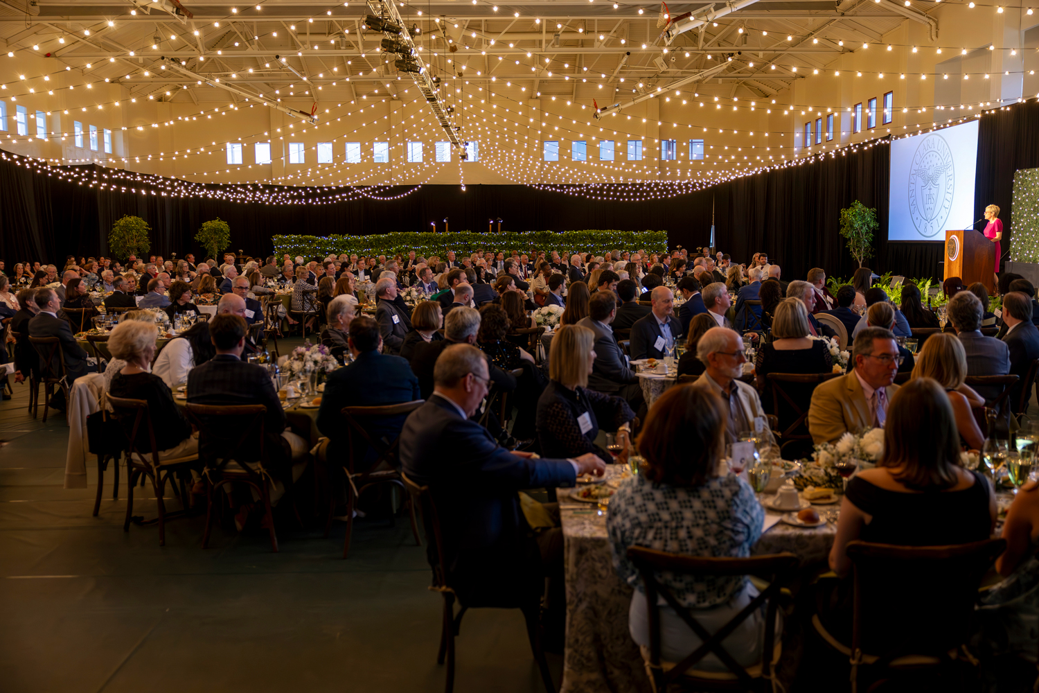 A large crowd seated at many tables pays attention to a speaker at a podium