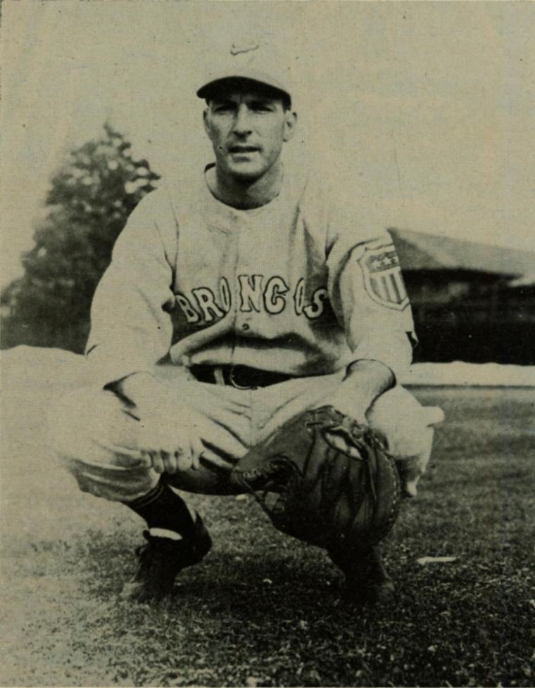 A sepia photo of a baseball player in uniform, squatting on a field.