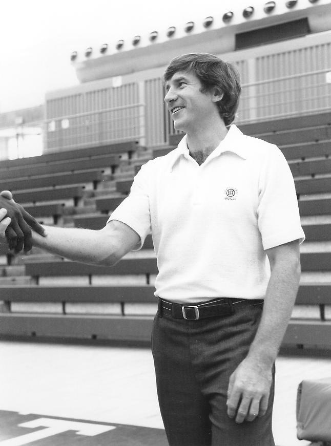 A person smiling in a sports stadium, shaking hands.