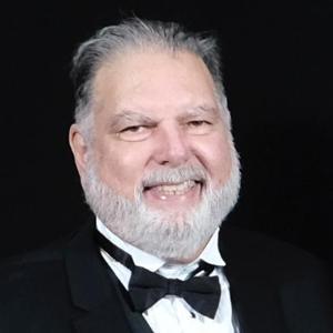 A man in a tuxedo smiling against a black background.