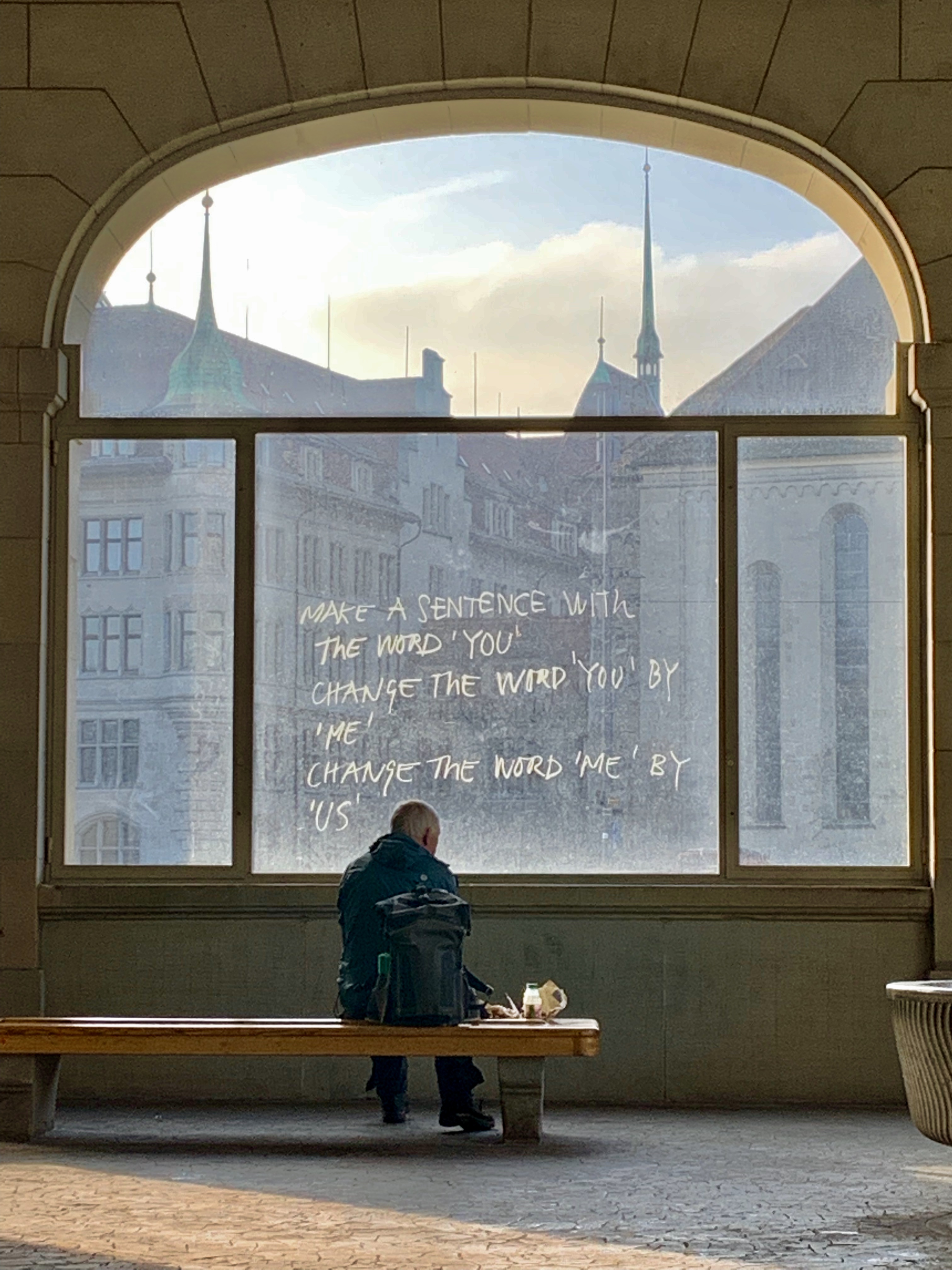 Person sitting on a bench inside a building with arched windows displaying city view.
