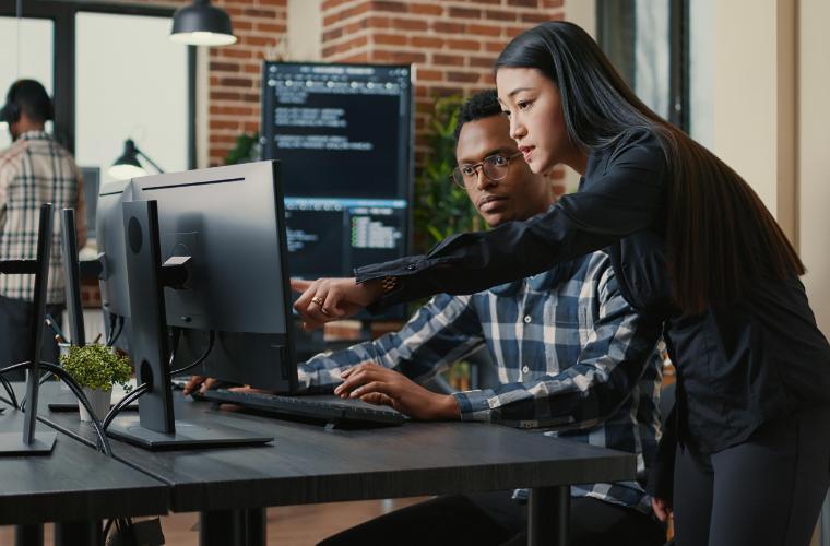 Man sitting in front of a computer, while a woman next to him points to the computer. 