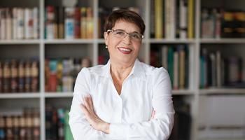 A mature female professor stands smiling in front of a bookcase 