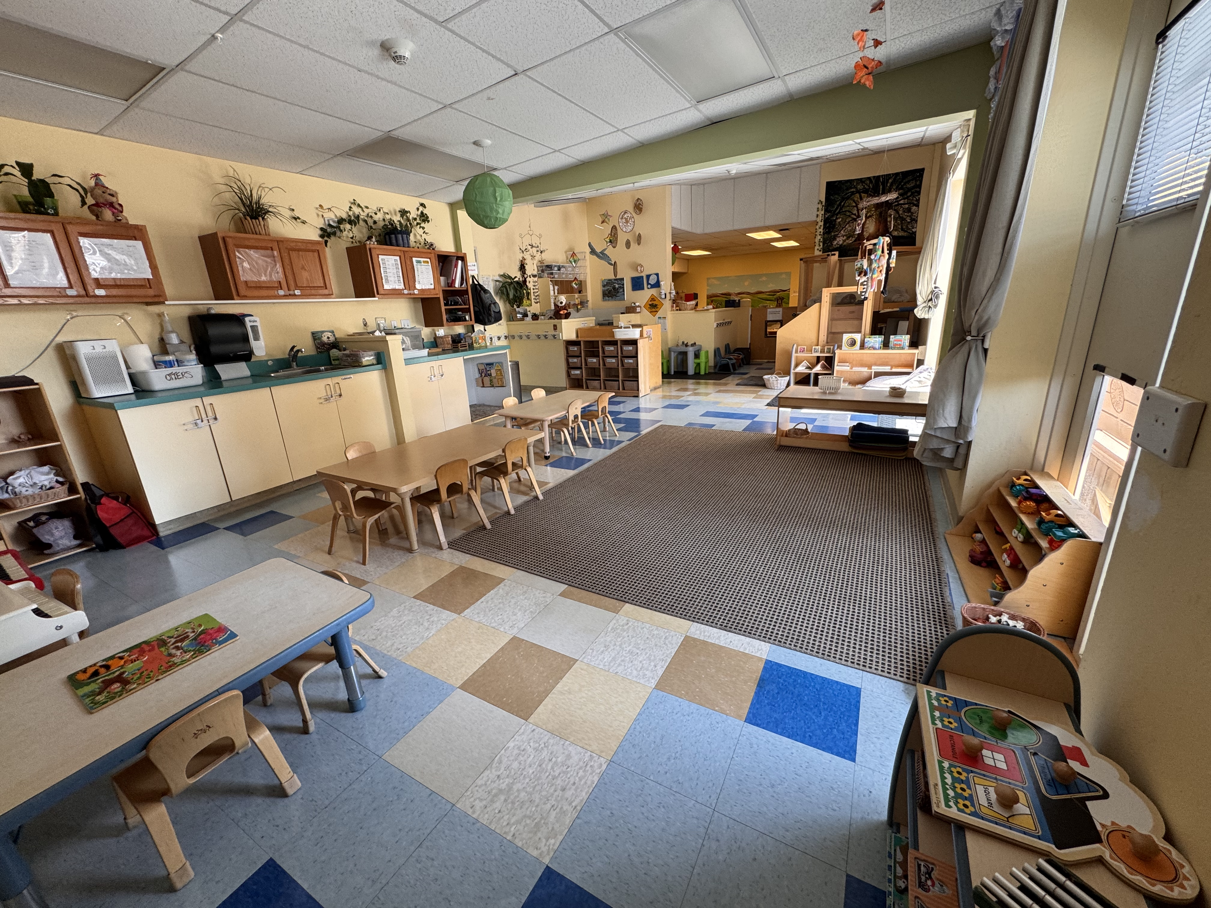 View of the otter classroom from the entrance to the storage locker, you can see cabinets with teaching supplies, the circle time carpet open area, and tables with chairs for toddlers