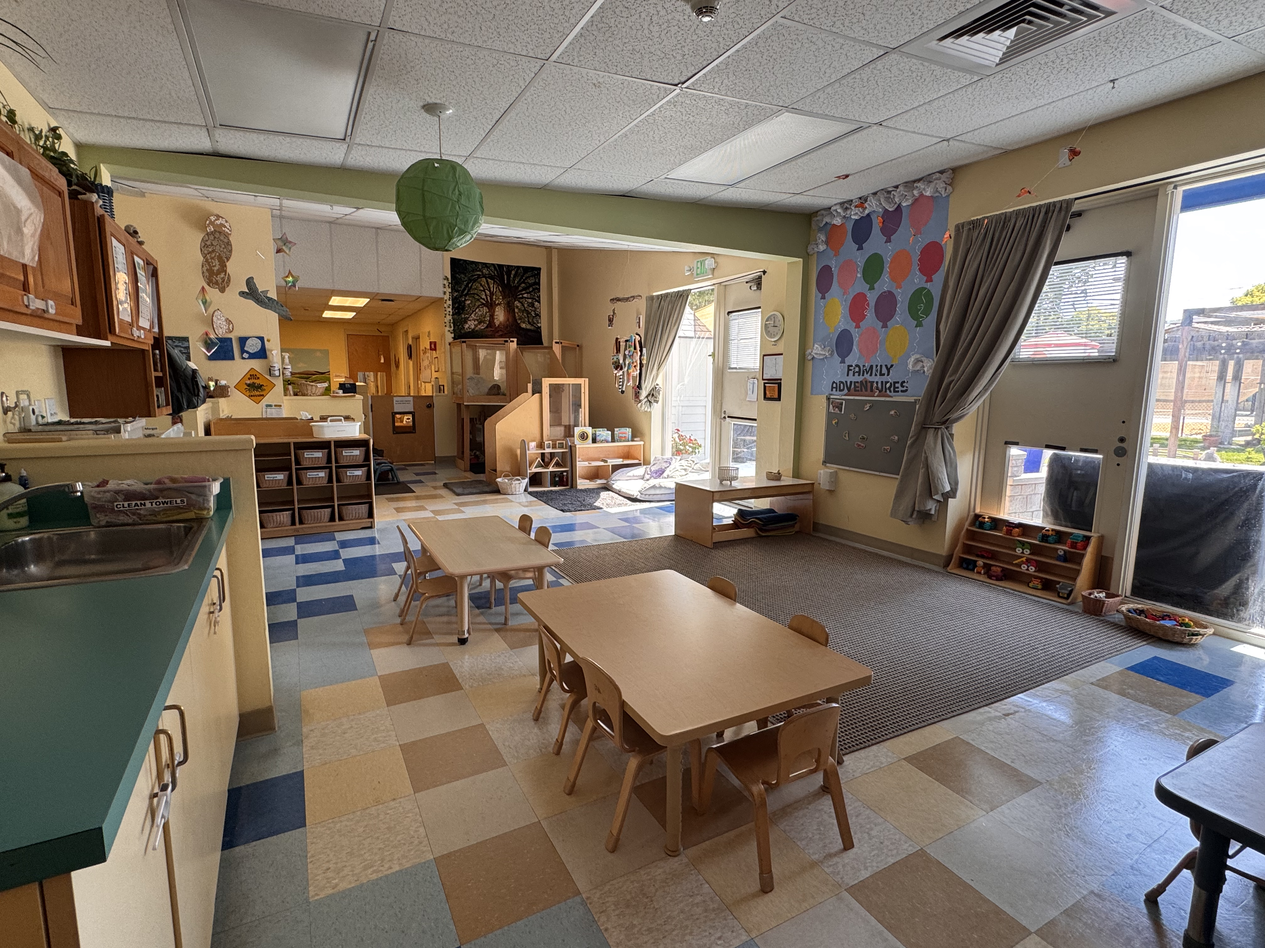 View of the otter classroom from the storage cabinets, you see part of the storage cabinets, circle time area, tables and chairs, in the distance the cubbies for childrens supplies, and the toddler jungle gym and entrance to the starfish room 