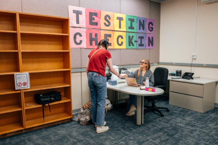 A staff member under a Testing Check-in sign hands over a piece of paper to a student. The students bag is deposited onto an empty bookshelf.