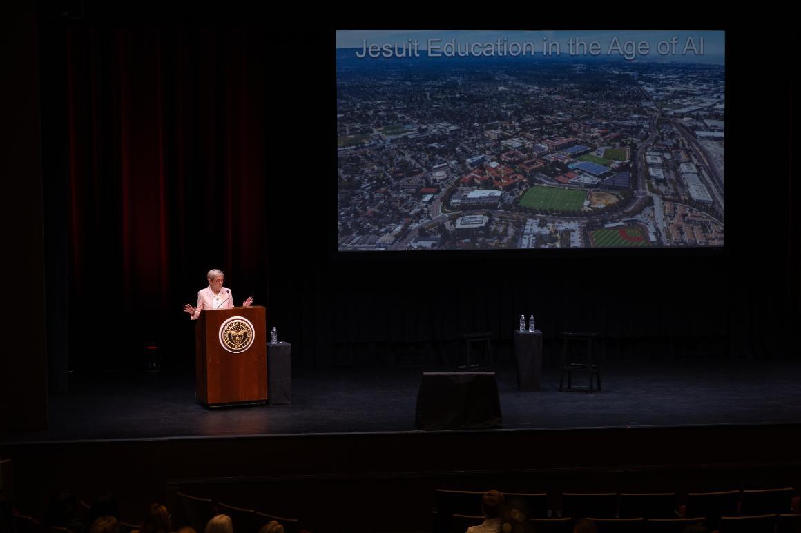 SCU President Julie Sullivan gestures while speaking at a podium beneath a slide titled “Jesuit Education in the Age of AI,” featuring an aerial view of campus.