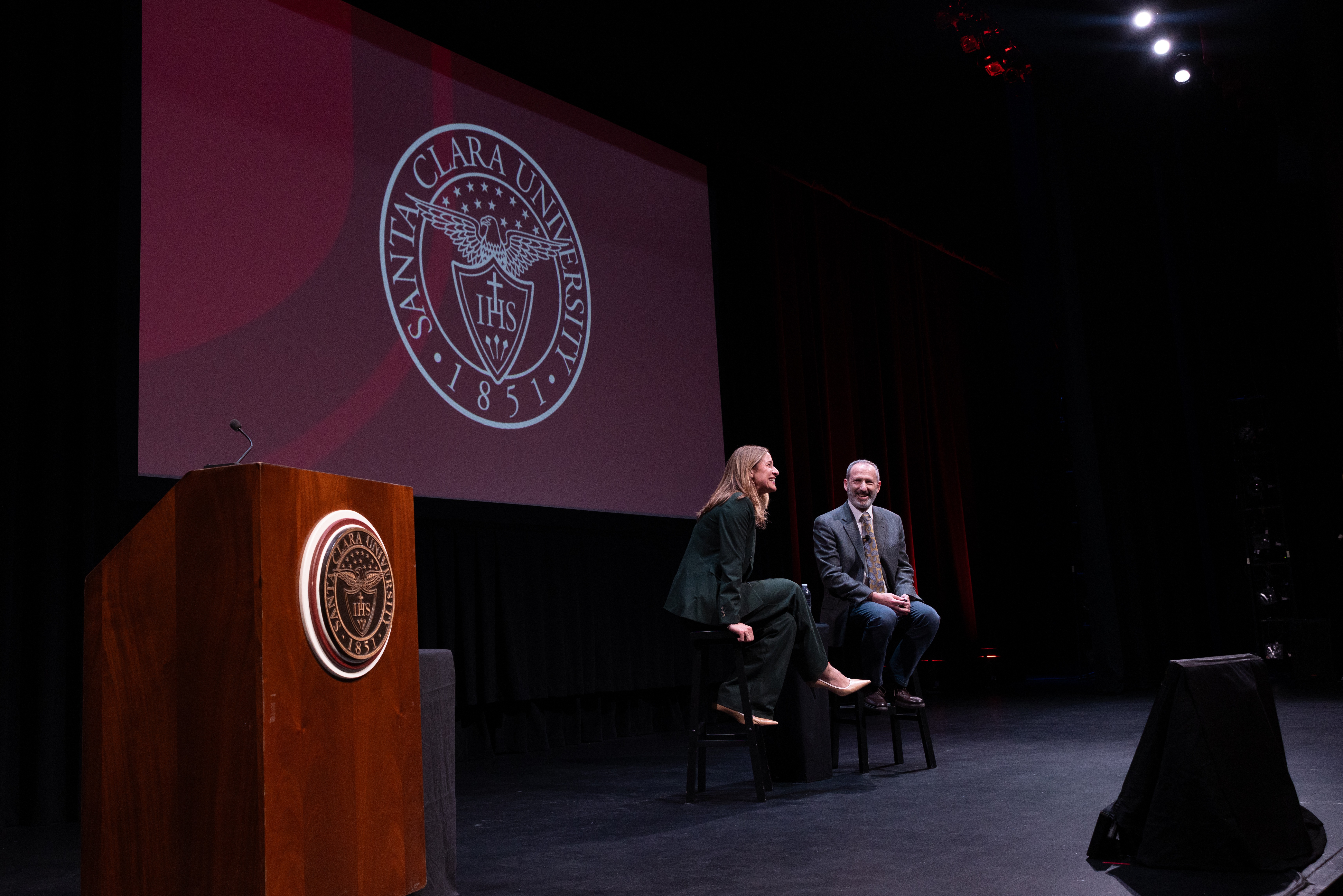 SCU Provost Jim Glaser and Lindsay Mazotti, planning dean for the Stevens School of Medicine and chief academic officer at Sutter Health, smile while seated onstage beneath the Santa Clara University seal. SCU Provost Jim Glaser and Lindsay Mazotti, planning dean for the Stevens School of Medicine and chief academic officer at Sutter Health, smile while seated onstage beneath the Santa Clara University seal.