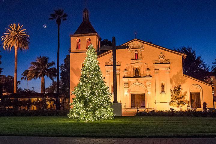 Night photo of lit-up Mission w/ lit Xmas tree in front.