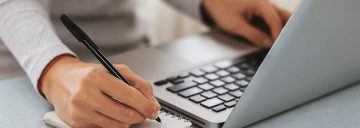 A person taking notes with a pen while using a laptop.