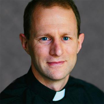 A man in a clerical shirt with a grey background.