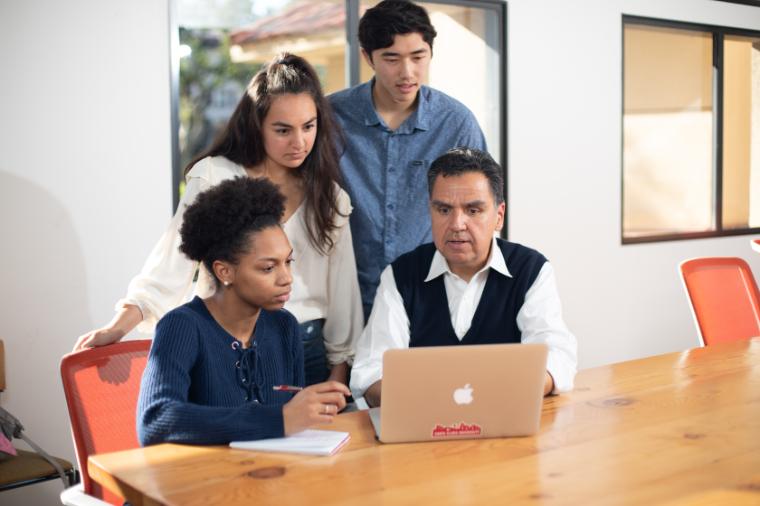 Faculty member in the classroom with students stand over a laptop