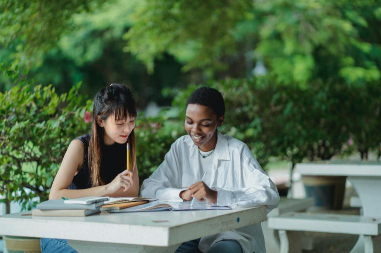 Two students working together outdoors at a table with books and papers. 