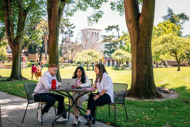 Three people sitting at a table with laptops outside