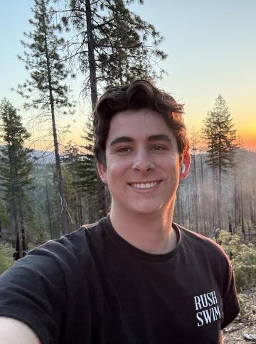 A smiling young man with dark wavy hair takes a selfie in a pine forest at sunset, wearing a black “RUSH SWIM” T-shirt.