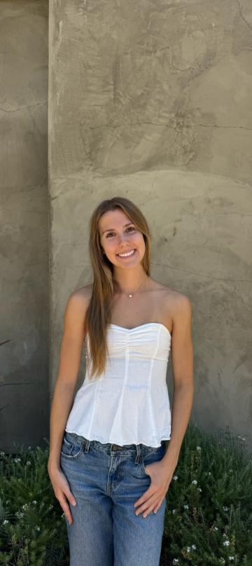 A smiling young woman with long light brown hair stands against a gray concrete wall in a white strapless top and jeans.