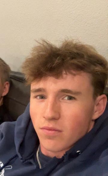 A close-up selfie of a young man with tousled light brown hair in a navy hoodie, in front of a plain wall.