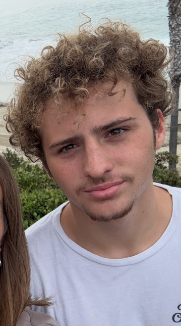 A close-up of a young man with curly sandy-brown hair and freckles in a white T-shirt, with the beach behind him.