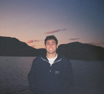 A smiling young man with short dark hair in a navy pullover stands by a lake at dusk, with mountains behind him.