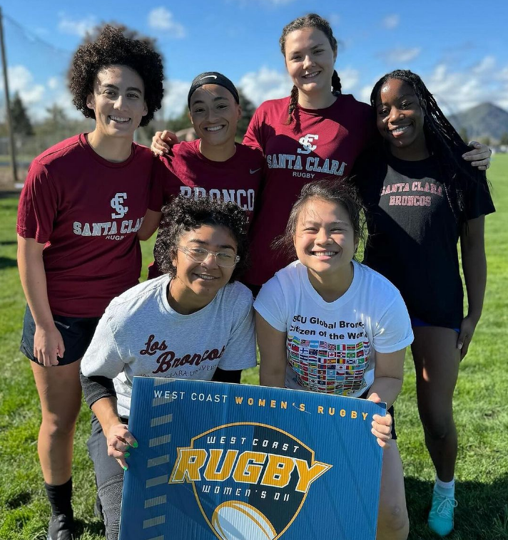 Picture shows 6 women's rugby players smiling on a field with a sign that says west coast rugby.