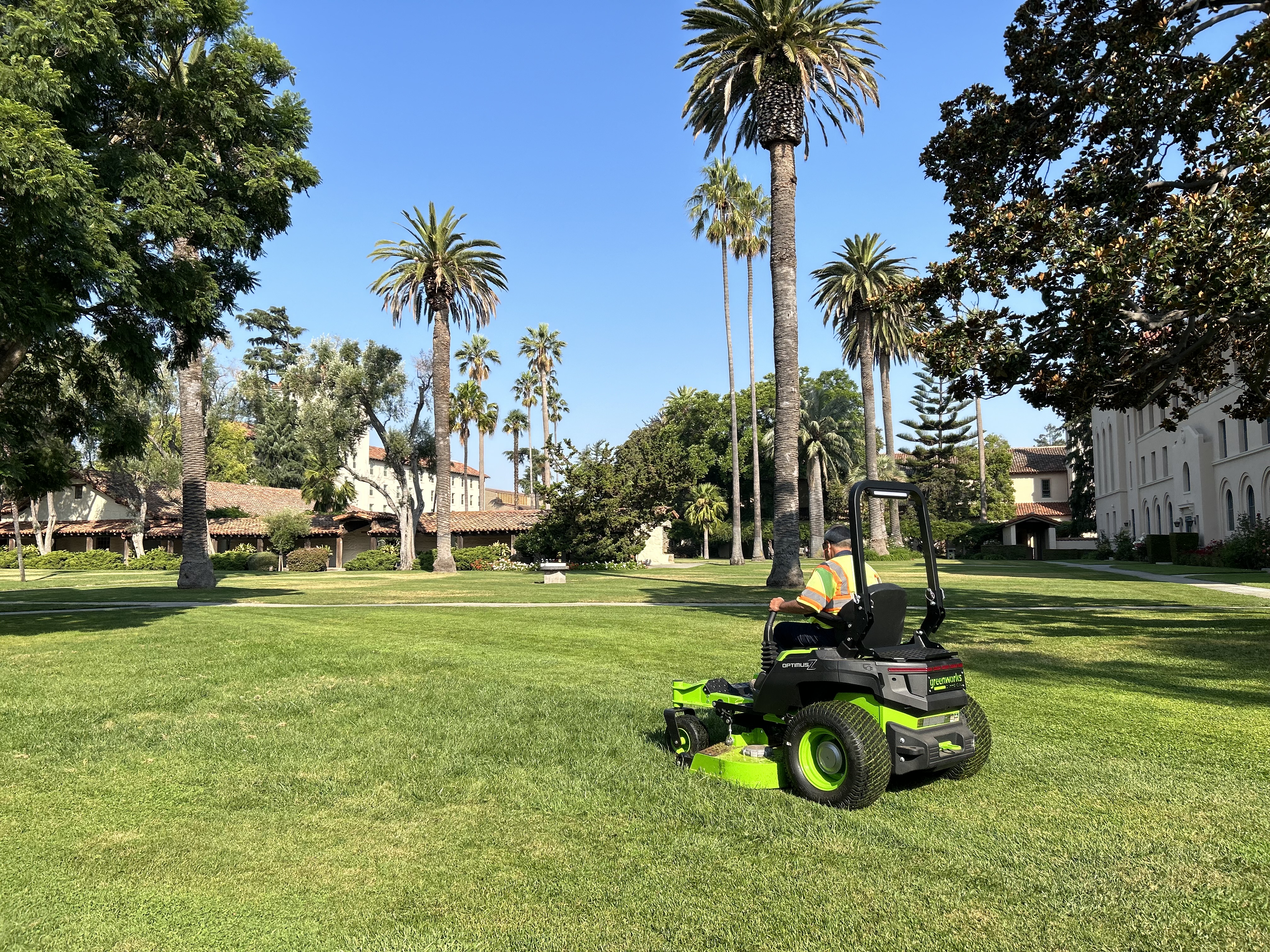 person on electric lawn mower in front of blue skies and palm trees on campus