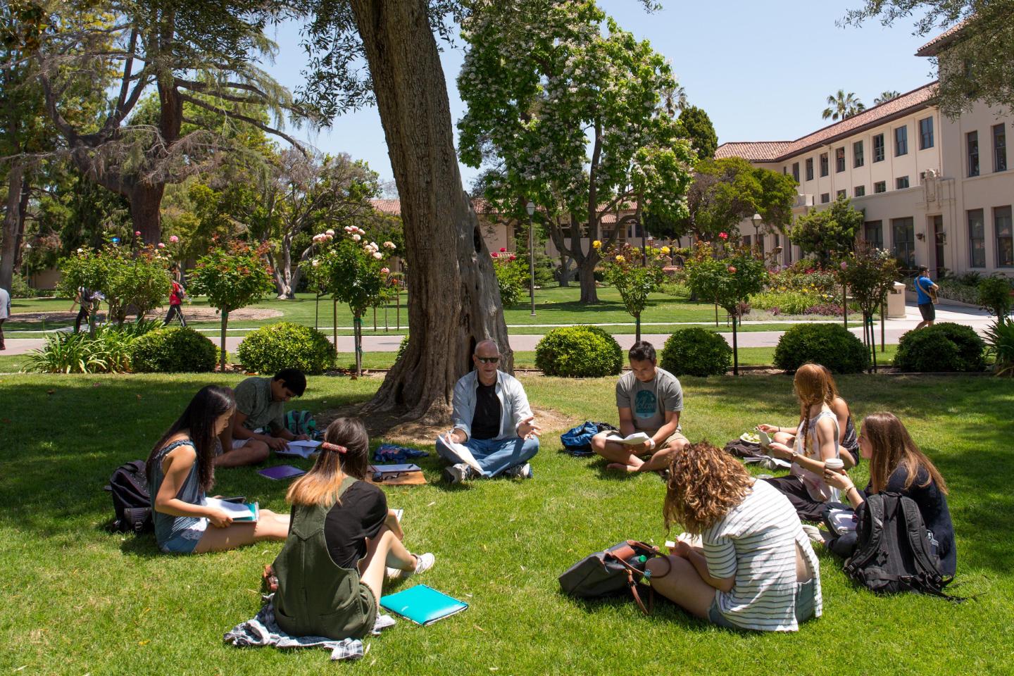 A faculty member and students sit outside in a circle discussing and taking notes