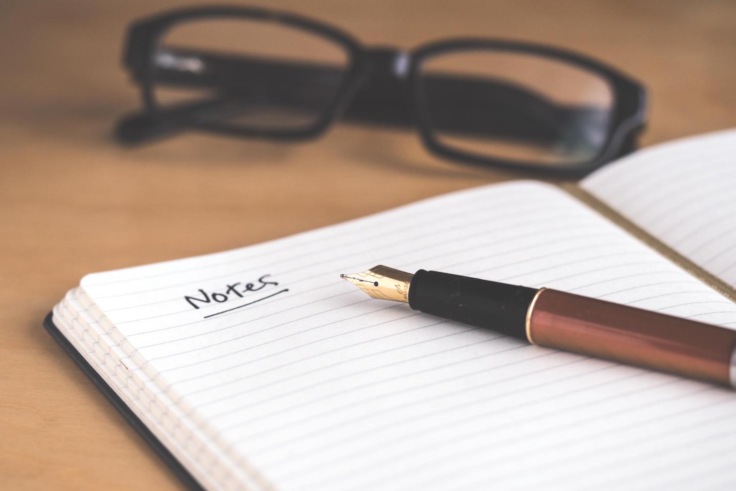 A fountain pen on an open notebook with glasses in the background