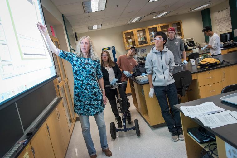 A faculty member points to the board in a lab while four students stand listening