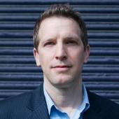 Alt text: Close-up of a man in a suit jacket against a dark striped background.