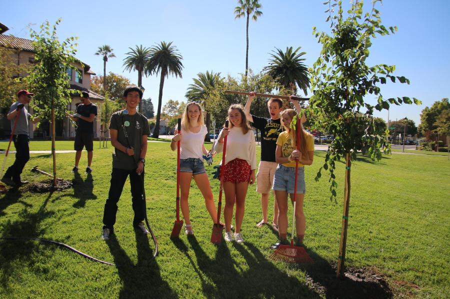 Five students smiling and holding shovels next to newly planted trees