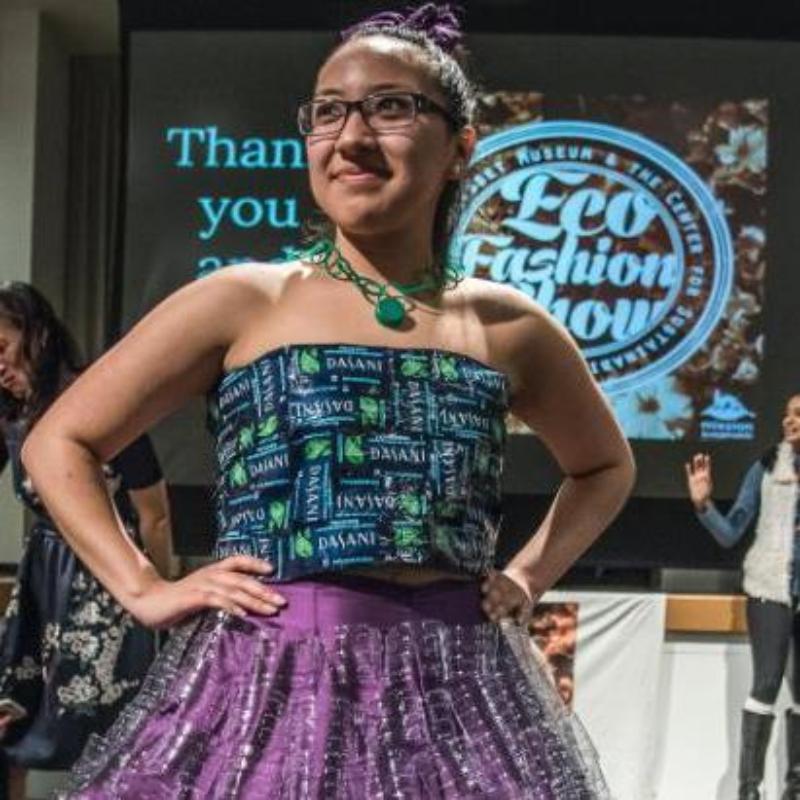 A student wearing a dress made of plastic bottles poses in front of an Eco Fashion Show sign