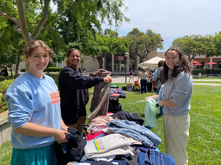 Three smiling students shopping at a table of clothes outside