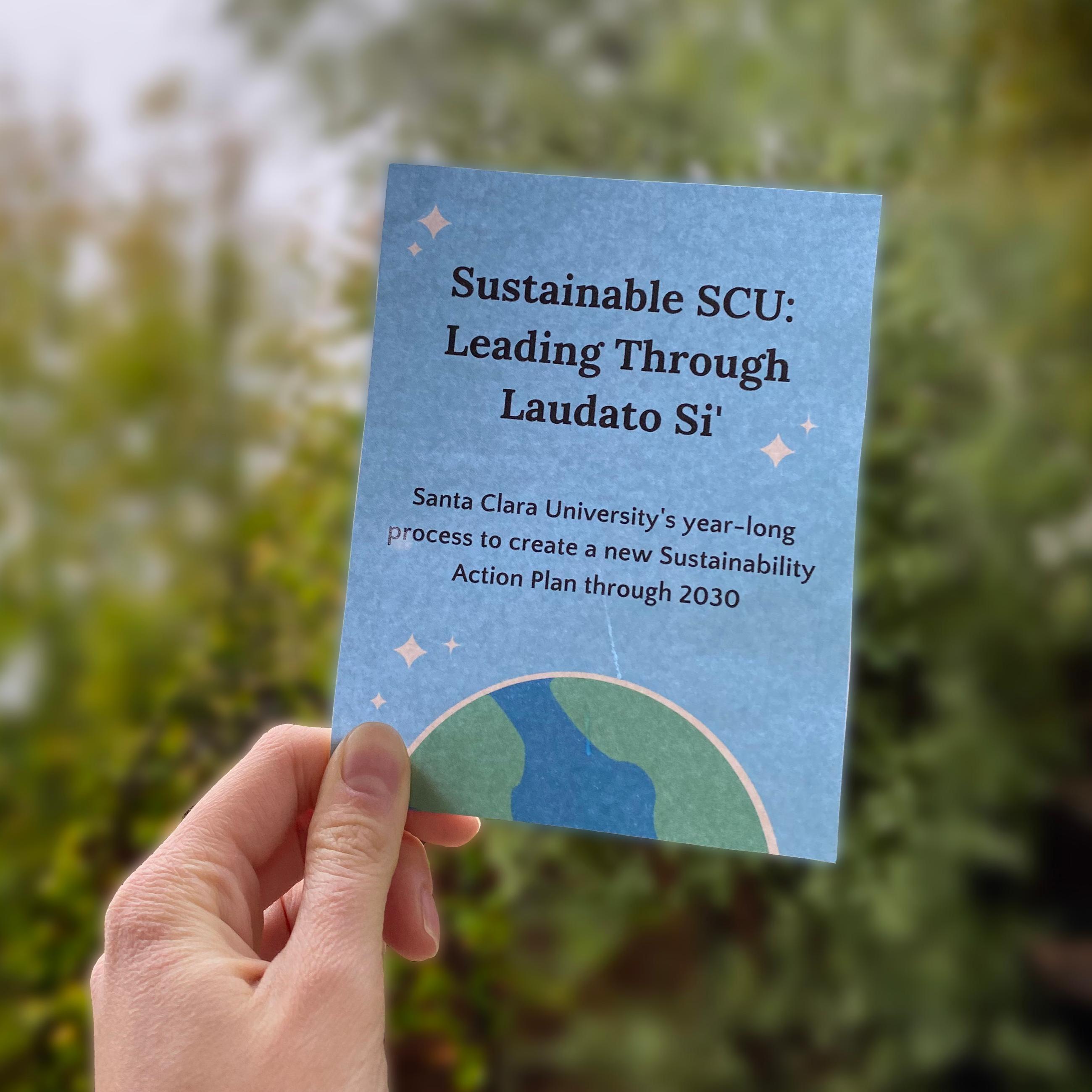 A hand holding up a card about Leading Through Laudato Si against a background of trees