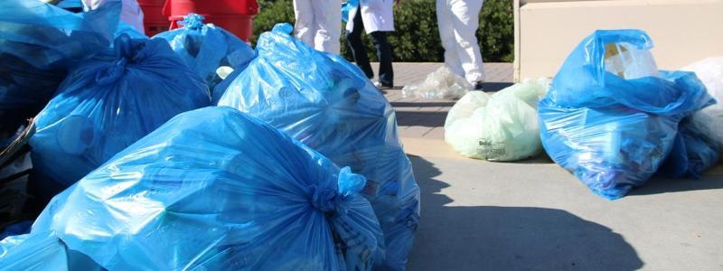 Several full blue recycling bags on the ground outside with students and a dumpster in the background 