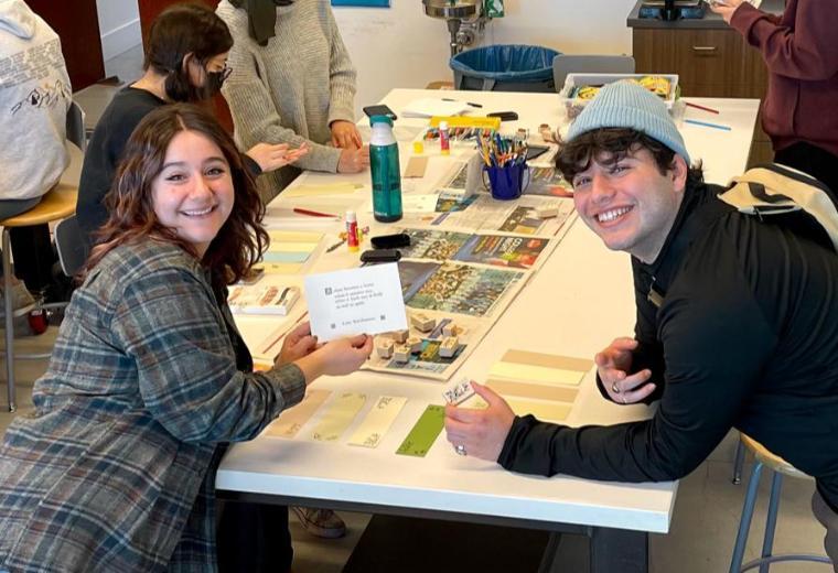 Two students in a busy work room smiling and holding up craft supplies and a letterpress card