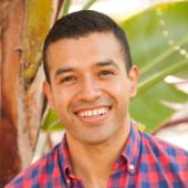 A man in a checkered shirt, smiling outdoors near palm leaves.