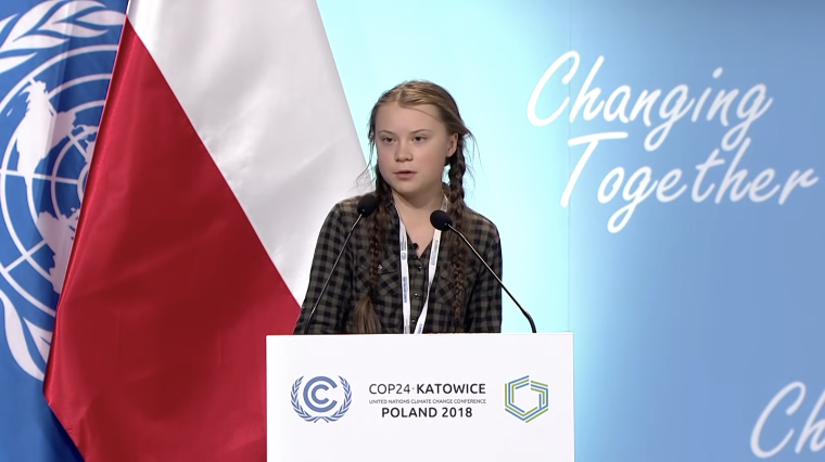 Climate activist Greta Thunberg speaks behind a podium at the UN COP24 Conference with flags of Poland and the UN in the background
