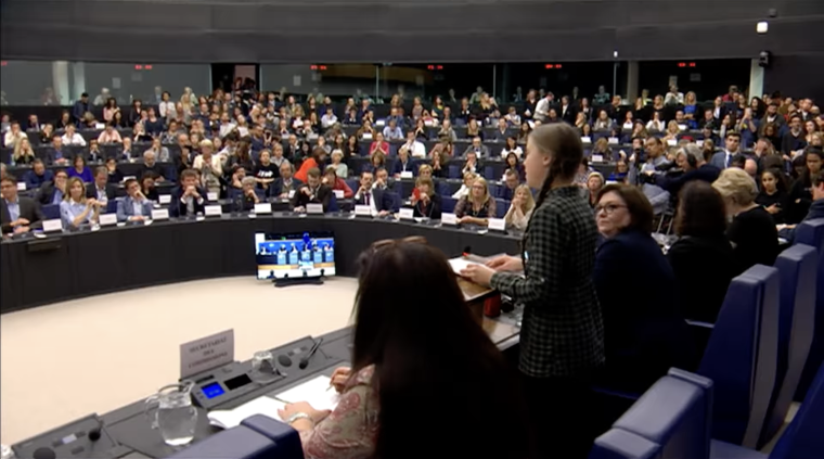 Climate activist Greta Thunberg speaks behind a podium facing a large assembly of members of the European Parliament
