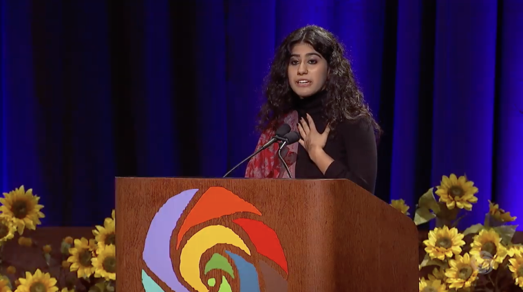 Pakistani immigrant and youth climate activist Mishka Banuri speaks at a podium with sunflowers on stage behind her