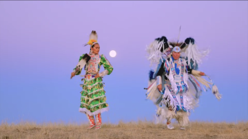 Artists Supaman and Acosia Red Elk dancing outside with a sunset in the background