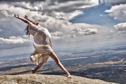 Dancer Camille Hanson poses on elevated ground overlooking a vast landscape with her arms in the air and back arched with the wind