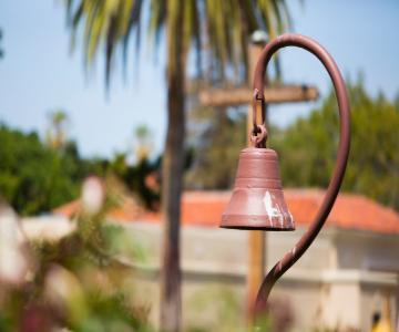 A bell hanging outdoors with palm trees and a building in the background.